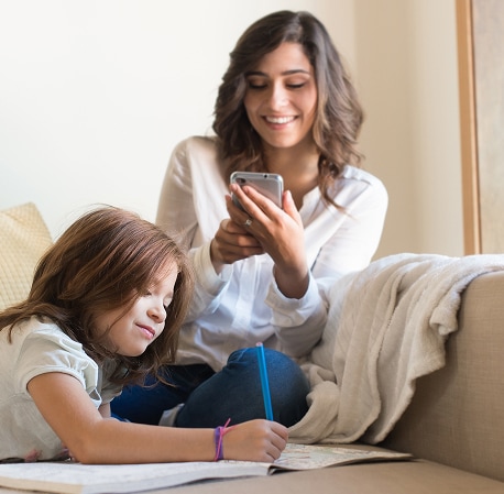A mother applying for private school on her phone while her daughter writes in a workbook